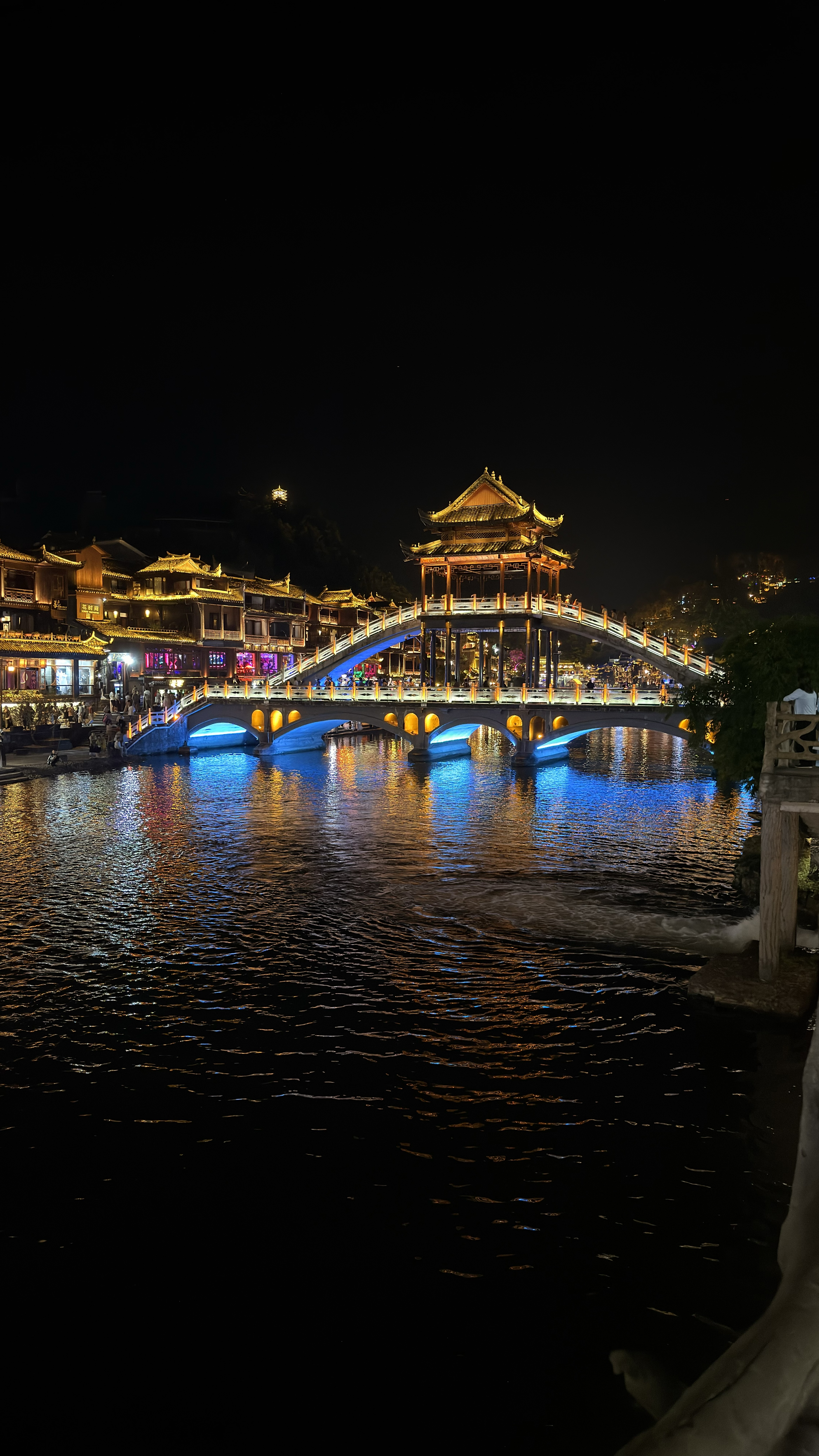 Bridge with traditional pavilion illuminated in blue at night in Fenghuang with reflection on the river