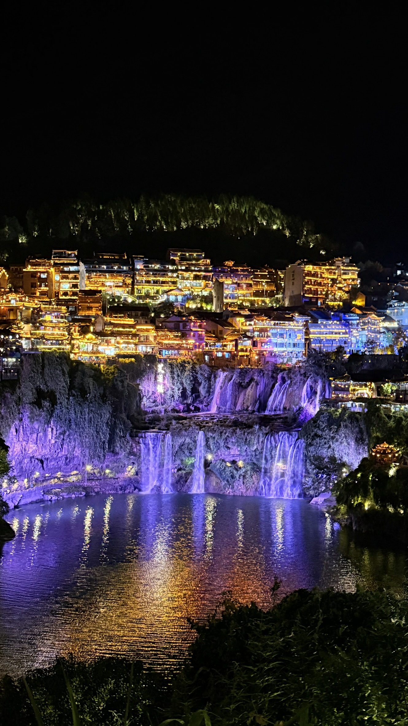 Vista panorâmica noturna de Furong Ancient Town com cachoeira iluminada em roxo e construções tradicionais
